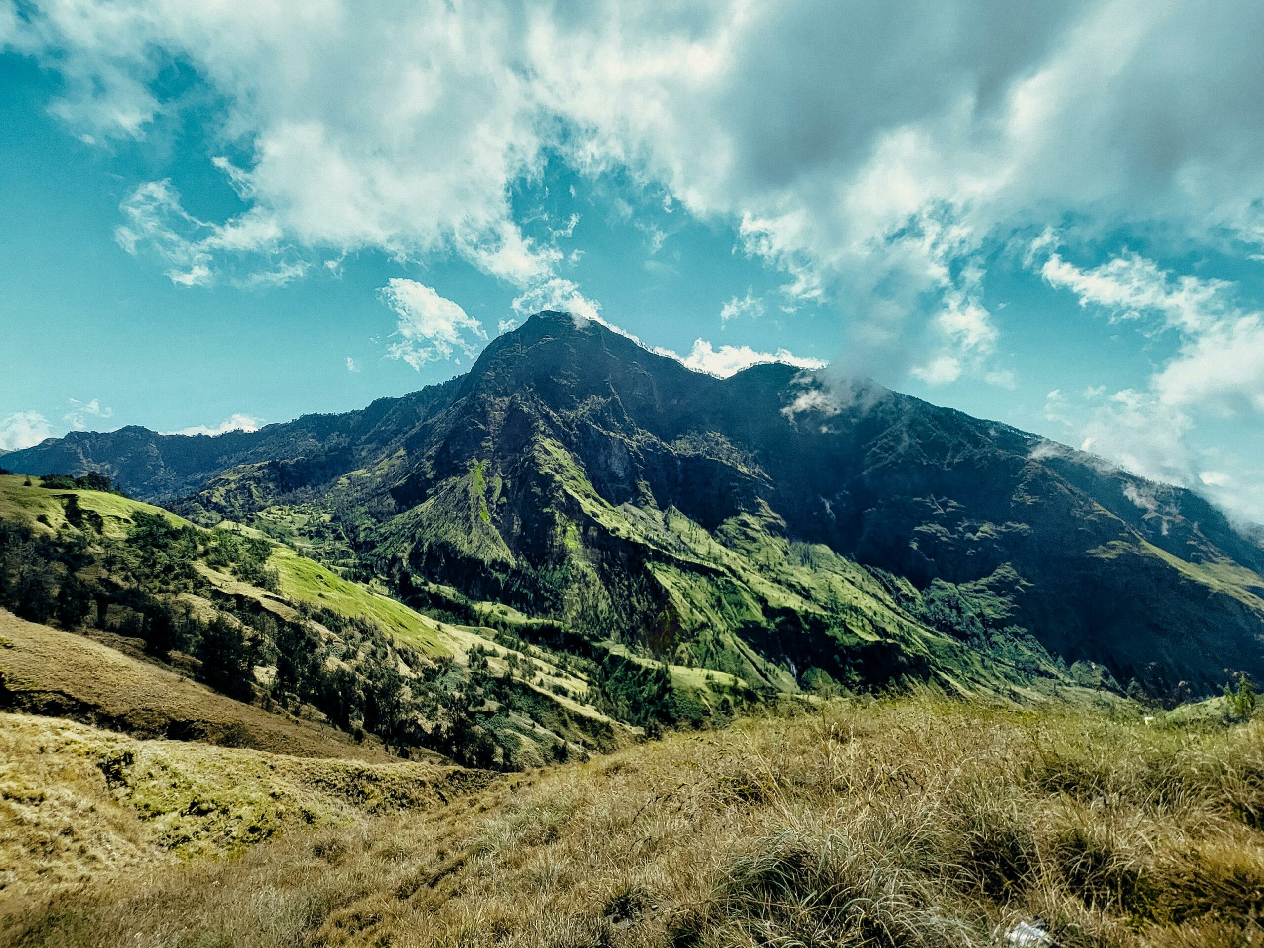 Vibrant view of Mount Rinjani, lush green valleys, and cloud-filled sky in Indonesia.
